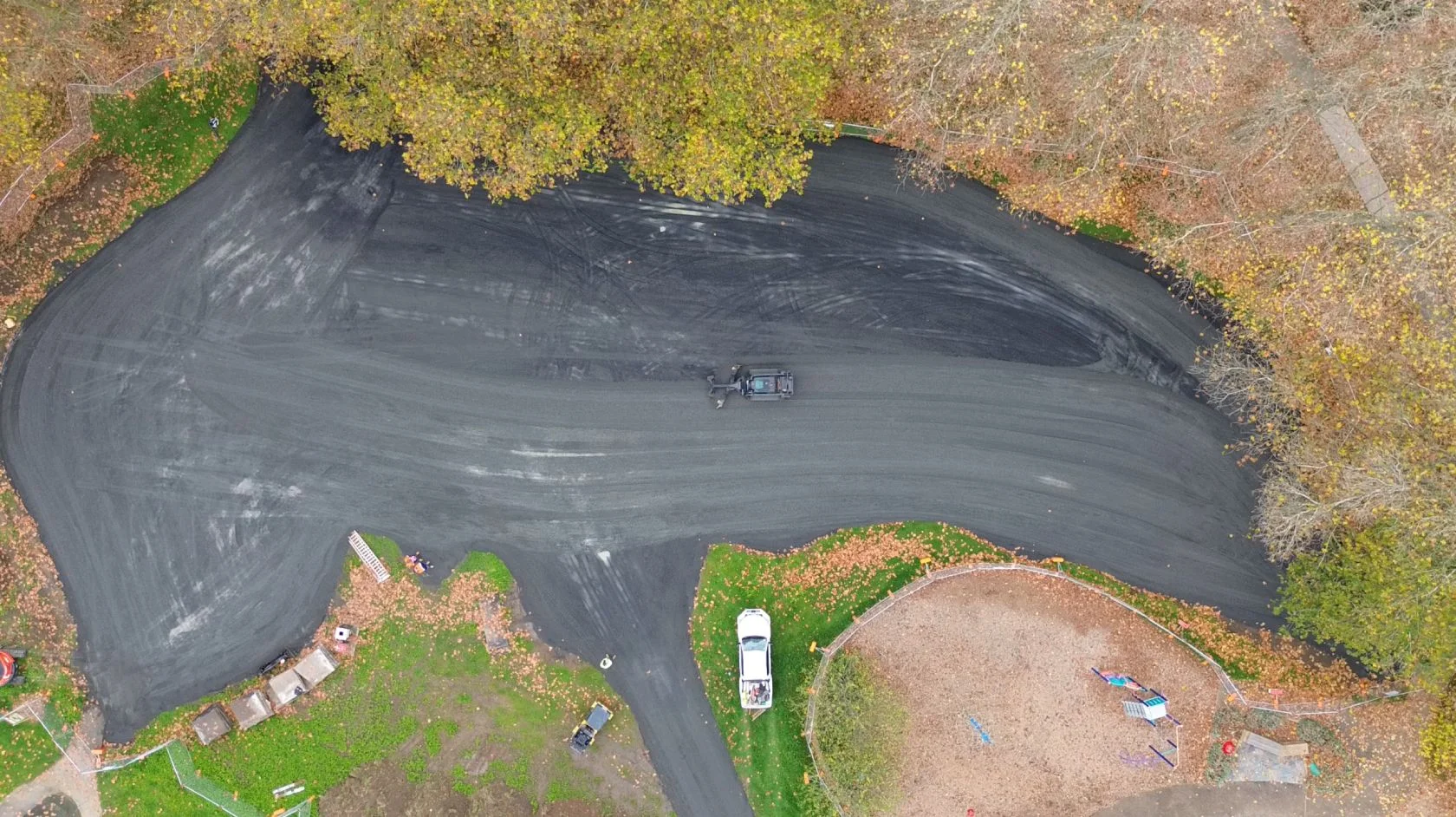 Aerial view of a newly paved road bordered by grass and trees, with construction vehicles, autumn leaves, and a playground in the lower right corner.