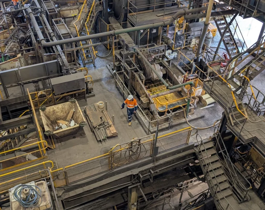 Aerial view of a large industrial factory with machinery, metal structures, and three workers in safety gear and orange vests performing tasks on different levels.
