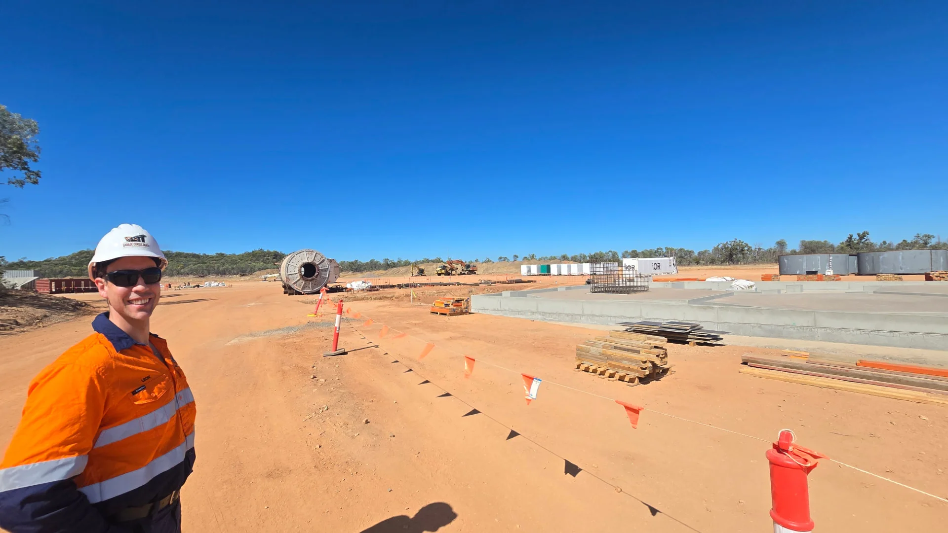 A construction worker in orange safety gear stands at a dusty construction site with concrete foundations, building materials, and equipment under a clear blue sky.