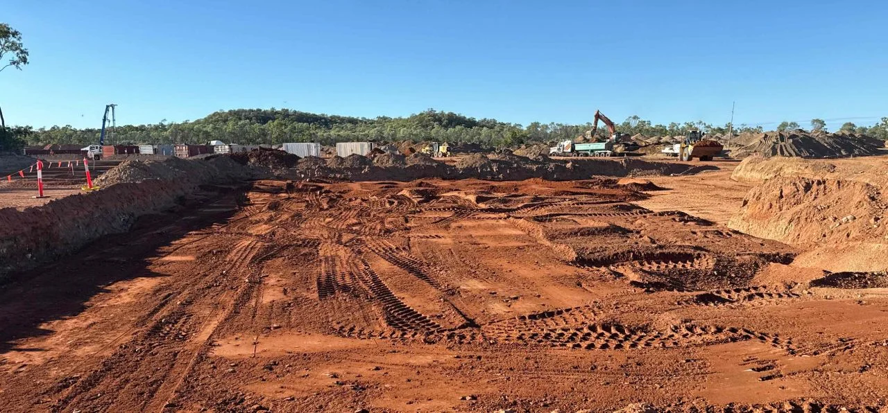 A construction site with red dirt, tire tracks, mounds of earth, and construction equipment in the background under a clear blue sky. Trees and containers line the edge of the site.