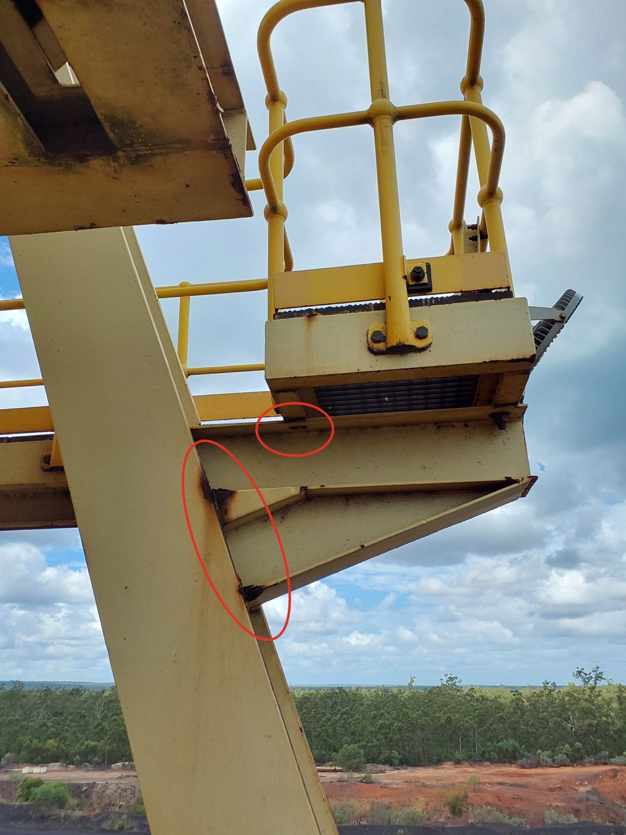 A metal platform with yellow railings is shown against a cloudy sky. Two red circles highlight structural cracks or damage on the support beam under the platform. Trees and a reddish ground are visible below.