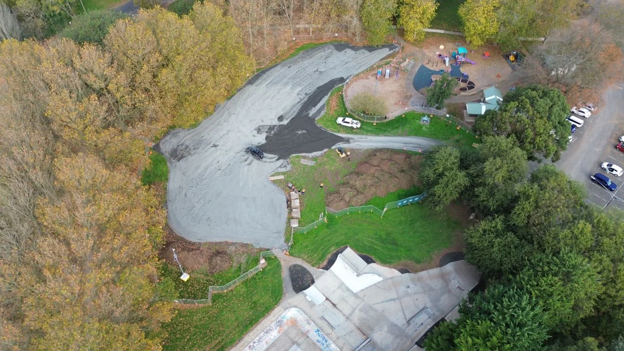 Aerial view of a park with trees, a playground, a parking area, and a partially paved section under construction near finished concrete paths and green grassy areas.