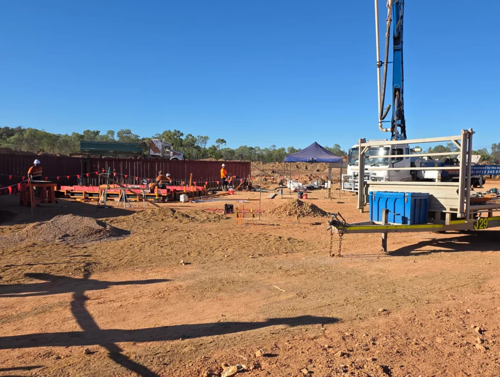 Construction site with workers wearing safety gear, equipment, and materials scattered around. A trailer and blue tent are visible, with trees and a clear sky in the background.