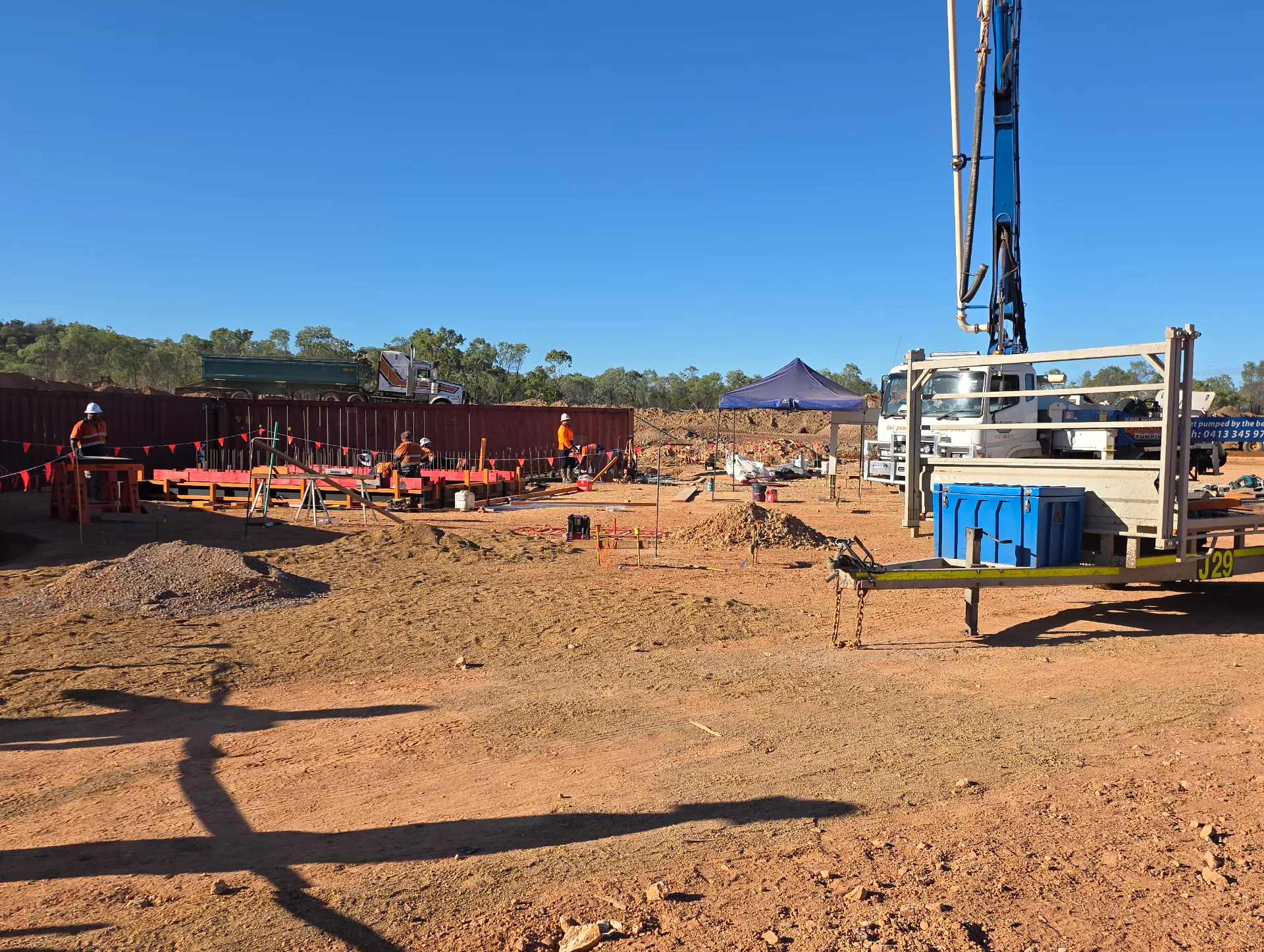Construction site with workers wearing safety gear, equipment, and materials scattered around. A trailer and blue tent are visible, with trees and a clear sky in the background.