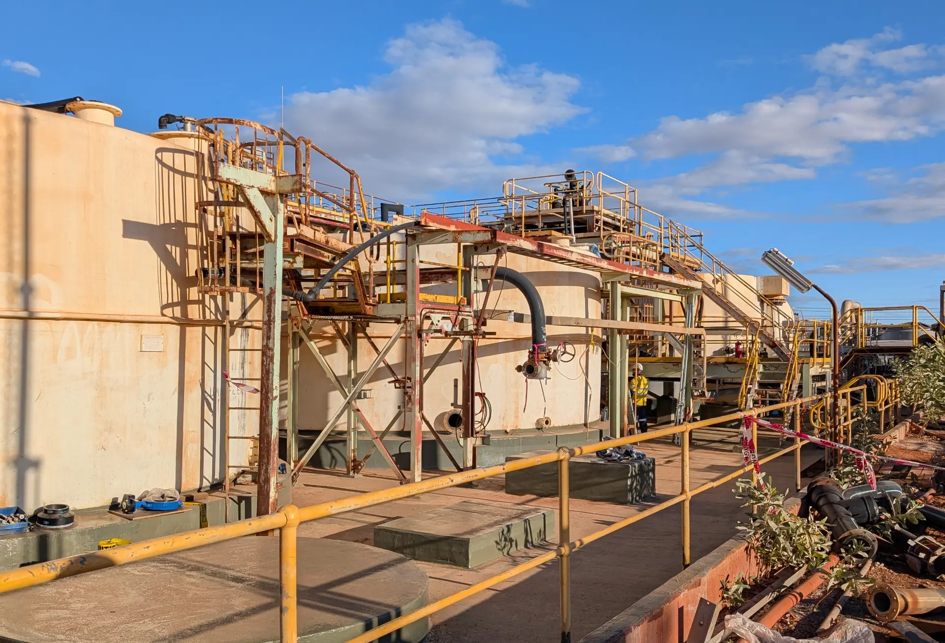 Large industrial tanks with yellow railings, ladders, and pipes under a blue sky with clouds. Some machinery and equipment are scattered around the site. Vegetation is visible on the right side of the image.
