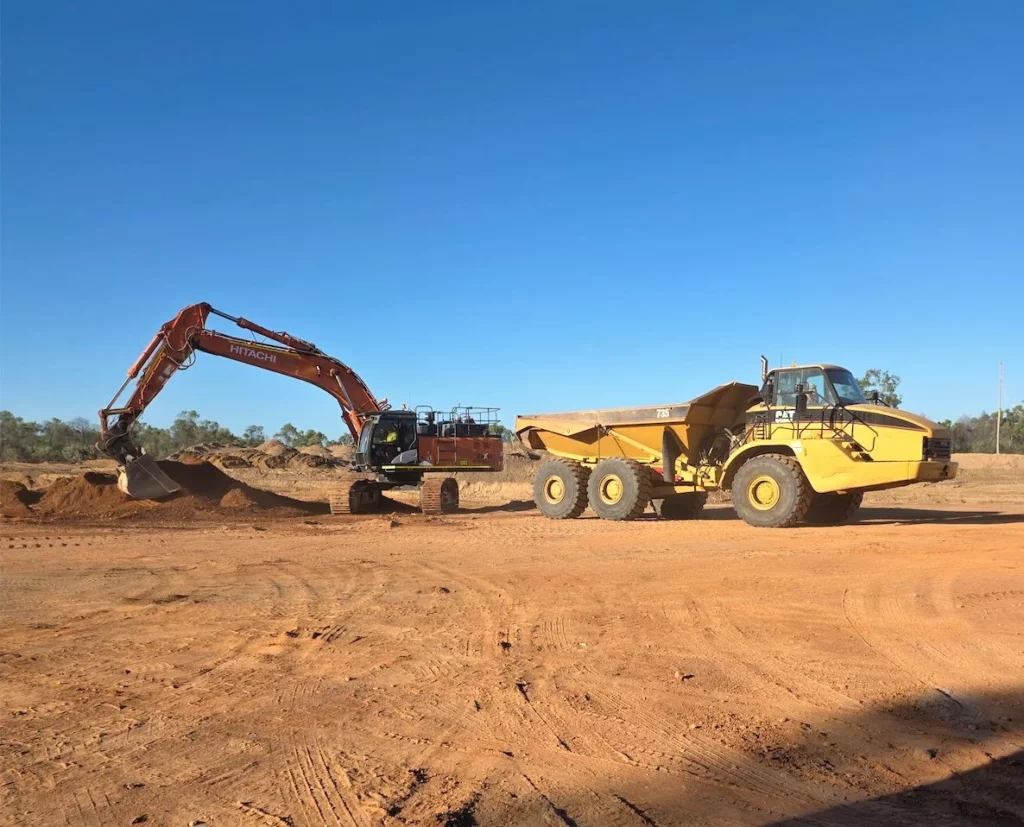 An excavator loads dirt into a large yellow dump truck at a construction site under a clear blue sky.